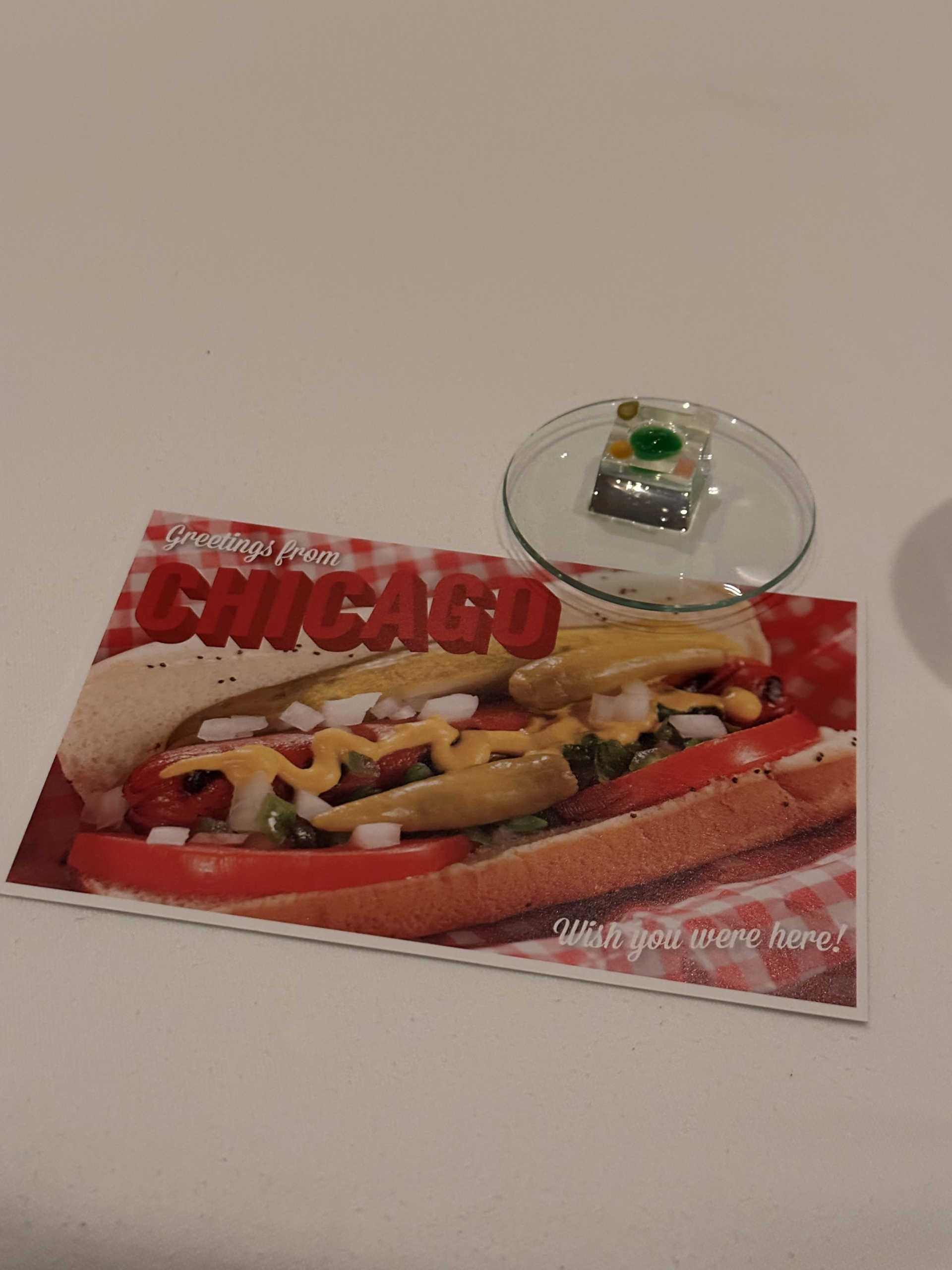 A tiny edible course on a glass dish beside a Greetings from Chicago postcard at Alinea